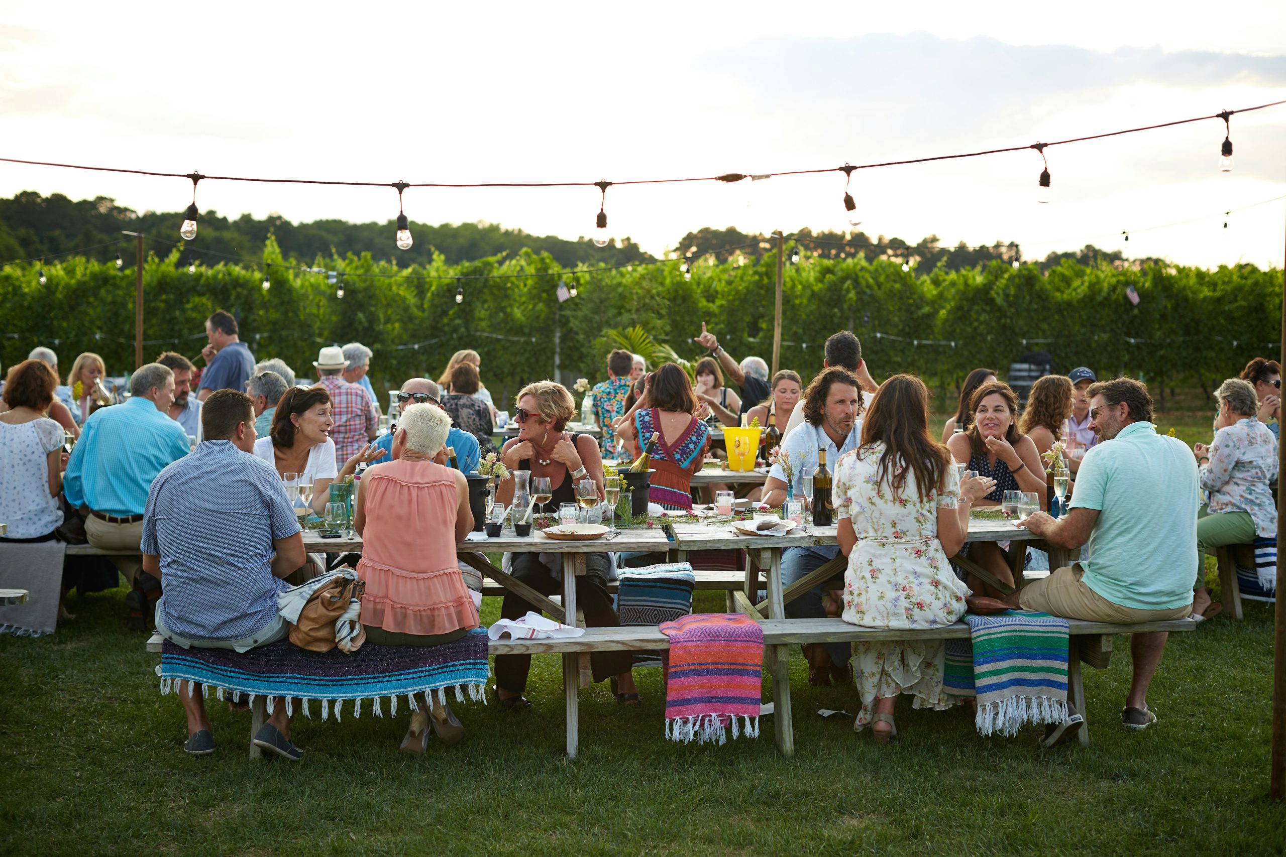 A large group of friends and family gathered outdoors at long tables beneath string lights at sunset, sharing food, laughter and memories at a living funeral celebration.
