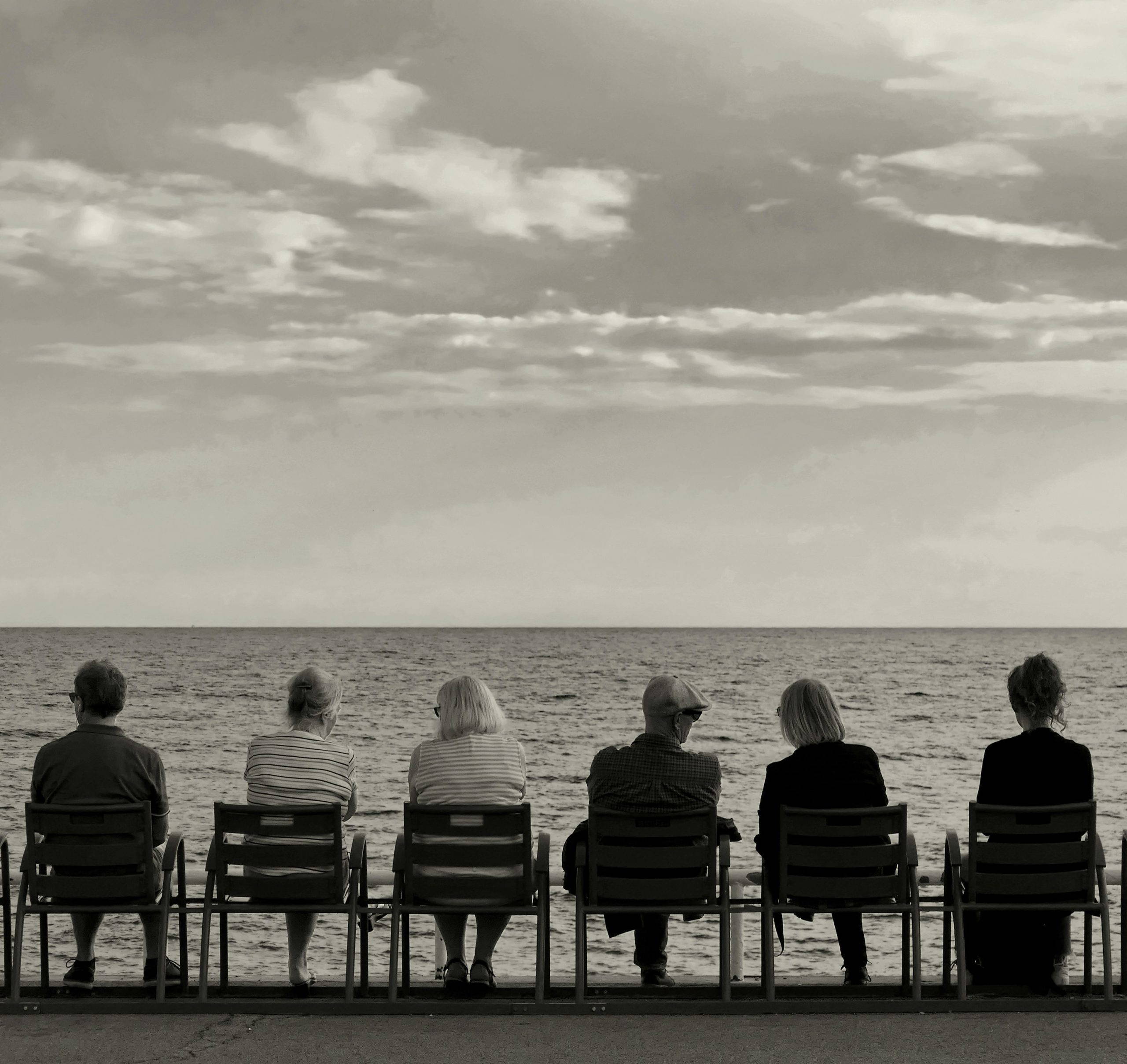A group of people seated together in chairs looking out to sea, sharing a quiet moment of reflection and remembrance at a memorial gathering.